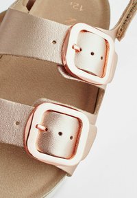 Metallisk roseguld sandal med två justerbara remmar med fyrkantiga metallspännen och en mjuk innerfoder, som framhäver en slät textur.