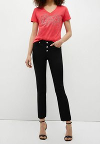 Camiseta roja de cuello en V con estampado plateado "Luv Jo". Jeans negros de tiro alto con cinco cierres de botón plateados. Sandalias de tacón negro con tiras.