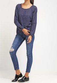 Marinblå långärmad tröja med cirkulärt mönster, kombinerad med blå tajta jeans med ett rivet knä. Glansiga svarta snörskor fulländar outfiten.