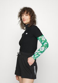 Top preto com textura em canelado de mangas curtas e gola alta, acompanhado por uma manga longa com padrão floral verde. Combinado com calções pretos de cordão com detalhe de logotipo.