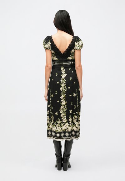 Zwarte bloemige jurk met korte pofmouwen, diepe V-rug, versierd met crème en groene bloemmotieven en rand aan de zoom. Feiten met laarzen zichtbaar.