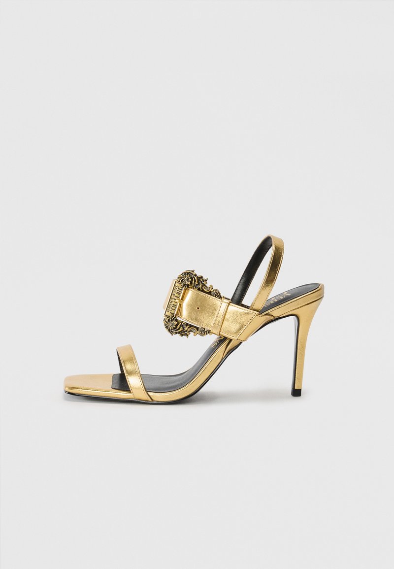 Auksinės metalinės aukštakulnės šlepetės su dekoratyvine sagtimi ir dviem siaurais dirželiais per pėdą. Juodas padas kontrastuoja su blizgiu viršumi.