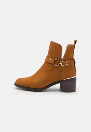 Brown suede ankle boot with a rounded toe, mid-height block heel, and a decorative adjustable strap featuring a circular buckle.