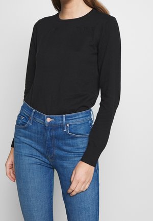 Mujer con una camisa negra de manga larga y jeans azul de talle alto, posando frente a un fondo blanco liso.
