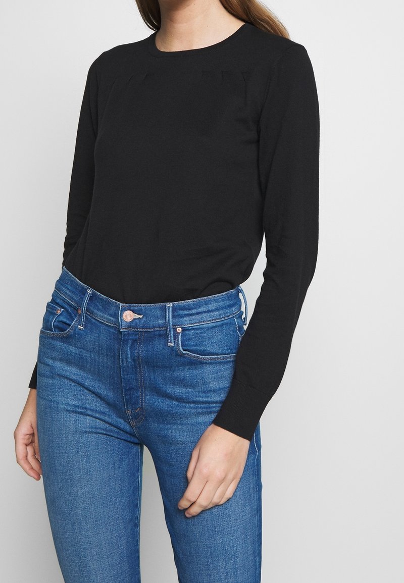 Femme portant une chemise noire à manches longues et un jean bleu taille haute, debout devant un fond blanc uni.