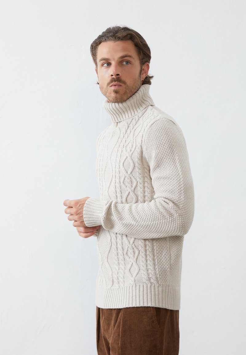 Beige cable knit turtleneck sweater with textured patterns and ribbed cuffs. Model is wearing brown corduroy pants, facing left.