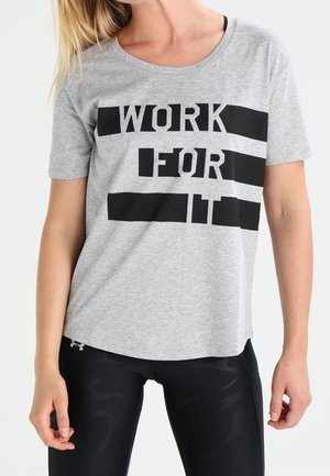 Femme portant un t-shirt gris avec texte noir "WORK FOR IT" et des leggings de sport noirs, debout les bras détendus le long du corps.