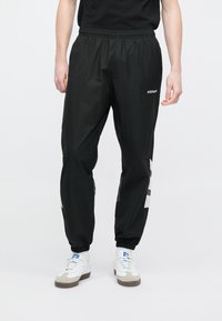 Muž oblečený v černých teplácích Adidas s bílým logem a bílými teniskami na jednoduchém bílém pozadí.