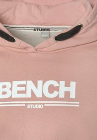 Sweat à capuche rose avec cordons noirs, comportant le texte "BENCH" en grandes lettres blanches et "STUDIO" en dessous en lettres blanches plus petites.