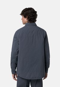Uomo con capelli ricci scuri che indossa una giacca a maniche lunghe grigia scura e pantaloni abbinati, rivolto di spalle su uno sfondo bianco.