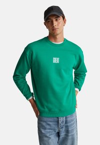 Jeune homme portant un sweat-shirt vert avec le texte "GREAT", un jean bleu clair, une casquette noire et une chemise blanche en dessous, se tenant avec les mains sur les hanches.