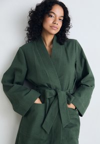 Robe vert kaki avec une surface texturée, des manches larges, une ceinture à nouer et des poches, présentée sur un mannequin devant un fond clair.