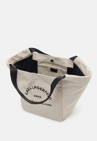 Beige canvas tygväska med svarta handtag, med tryckt text "KARL LAGERFELD PARIS" på ena sidan. Öppen topp och något slapp design.