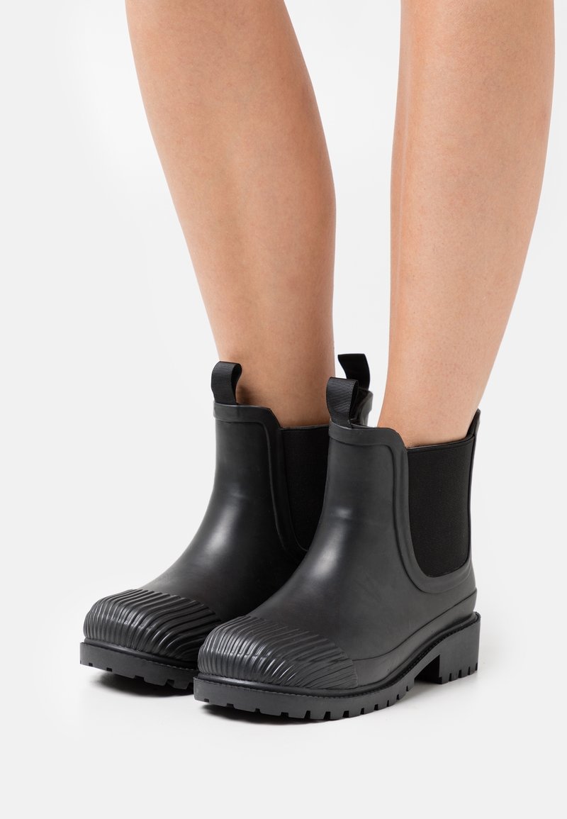 Bottes de cheville en caoutchouc noir avec des panneaux latéraux en élasthanne, un bout texturé et un petit talon bloc. Conçues pour le confort et la durabilité.