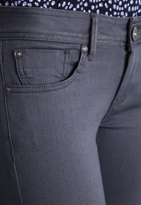 Un jean en denim gris foncé présente une coupe étroite, une poche avant avec des coutures décoratives et des accents en métal argenté près de la taille.