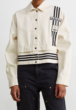 Person trägt eine kurze cremefarbene Adidas-Jacke mit schwarzen Streifen und Logos, kombiniert mit schwarzen Hosen mit sichtbaren Taschen und Nieten.