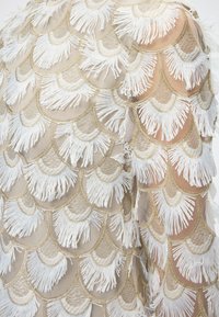 Διαφανές ύφασμα με επικαλυπτόμενα σχήματα βεντάλιας με χρυσά περιγράμματα, κεντημένα με λευκές κρόσσια που δημιουργούν υφασμένους κυματισμούς.