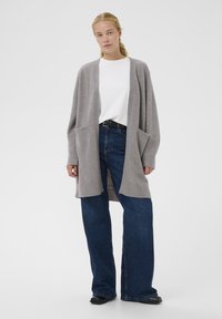 Cardigan lungo grigio realizzato in morbido tessuto a maglia, con vestibilità rilassata, apertura frontale e due tasche laterali. Indossato sopra una maglietta bianca e jeans blu.