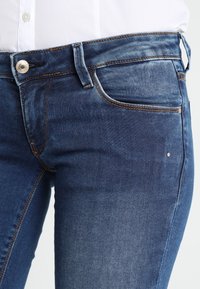 Jeans de mezclilla azul oscuro con corte recto, que presentan un diseño clásico de cinco bolsillos, un botón de metal y detalles de desgaste ligero.