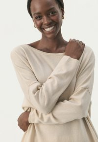 Beige Strickpullover mit einem schulterfreien Design, langen Ärmeln und gerippten Bündchen. Die weiche Textur und die lockere Passform betonen den Komfort.