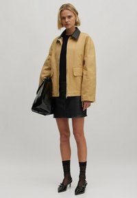 Beige oversized jas met een zwarte kraag, gecombineerd met een zwarte leren minirok en een glanzende zwarte handtas, gepresenteerd tegen een neutrale achtergrond.