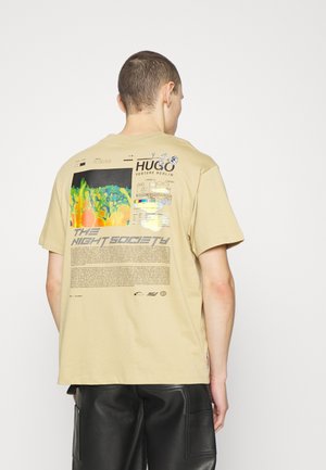 Eine Person trägt ein beiges T-Shirt mit grafischem Aufdruck und einem bunten abstrakten Design sowie dem Schriftzug "THE NIGHT SOCIETY" auf dem Rücken, kombiniert mit schwarzen Lederhosen.