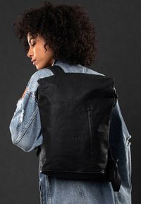Zaino a roll-top nero con texture liscia, tasca laterale con zip e spallacci regolabili, presentato su uno sfondo scuro.