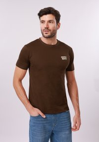 Homme portant un t-shirt marron "OSAKA" et un jean bleu, se tenant debout avec une main dans la poche, regardant légèrement sur le côté sur un fond uni.