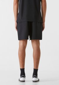 Schwarze Sportshorts mit elastischem Bund und Kordelzug. Getragen mit schwarzen Socken und weißen Turnschuhen, was eine lockere Passform zeigt.