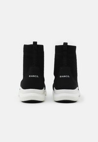 Schwarze gestrickte Knöchelsneaker mit einer weißen Sohle, ausgestattet mit einer Schlaufe zum Anziehen und einem Markenlabel auf der Rückseite, aus strukturiertem Stoff für zusätzlichen Komfort.