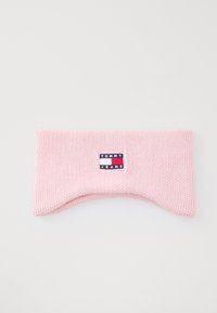 Ružičasti pleteni headband s zakrivljenim oblikom, koji ima istaknutu oznaku "TOMMY JEANS" u plavoj, bijeloj i crvenoj boji. Glatka tekstura.