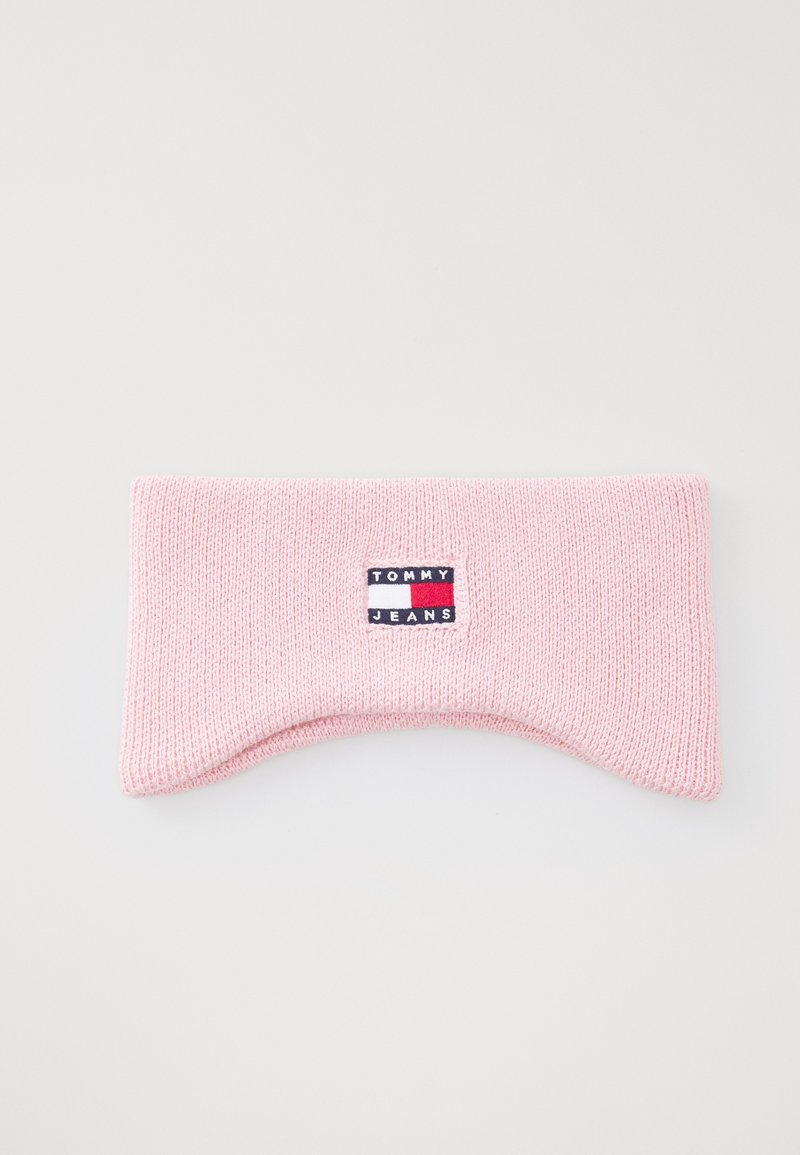 Ružičasti pleteni headband s zakrivljenim oblikom, koji ima istaknutu oznaku "TOMMY JEANS" u plavoj, bijeloj i crvenoj boji. Glatka tekstura.