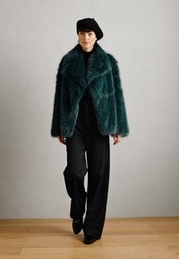 Tee-farbene Kunstpelzjacke mit breiten Revers, schwarzer Rollkragenpullover und schwarzen weiten Hosen; kombiniert mit einer schwarzen Baskenmütze und spitzen Schuhen.