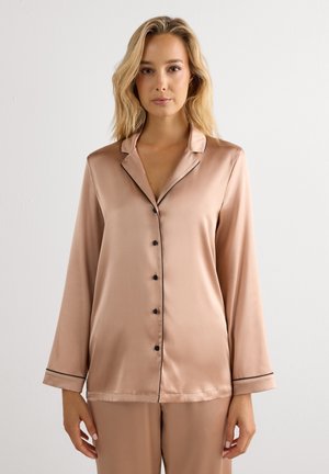 Frau mit blondem Haar, die ein beiges Satin-Pyjamaoberteil mit langen Ärmeln, Knopfleiste und schwarzer Paspel sowie passende Hosen trägt, vor einem schlichten hellen Hintergrund stehend.