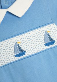 Lichtblauw polo-shirt met een witte kraag, voorzien van zigzag borduurwerk en twee blauwe zeilbootontwerpen in het midden.