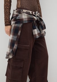 Braune Cargo-Hose mit Cargotaschen und einem karrierten Flanellhemd, das um die Taille gebunden ist und ein Muster in Blau, Braun und Creme aufweist.