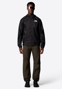 Veste coupe-vent noire avec une poche zippée, associée à un pantalon vert olive et des baskets noires. Lunettes de soleil avec des verres réfléchissants.