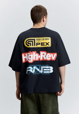Person trägt ein schwarzes T-Shirt mit bunten Schriftzügen "Apex", "High-Rev" und "A13" auf der Rückseite, kombiniert mit grünen Hosen.