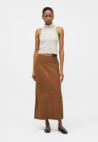 Γυναίκα όρθια, με ριγέ αμάνικη μπλούζα με ψηλό γιακά, μακριά καφέ φούστα και μαύρα φλατ παπούτσια, μπροστά σε απλό λευκό φόντο.