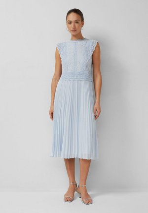 Vrouw die een mouwloze lichtblauwe kanten top en geplooide midi-rokjurk draagt met zilveren sandalen met hak, tegen een effen achtergrond.