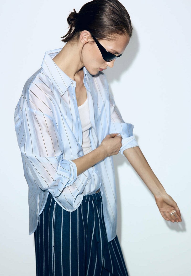 Mujer con gafas de sol negras estilo ojo de gato remangándose las mangas de una camisa azul claro a rayas, combinada con pantalones a rayas azul oscuro.