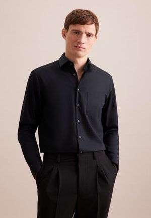 Ung mand iført en tætsiddende sort skjorte med knapper og brystlomme samt sorte plisserede bukser, hænderne i lommerne, neutral baggrund.