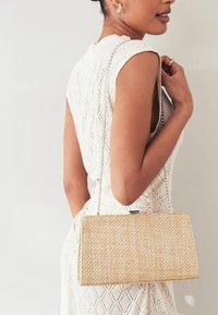 Mujer con un vestido blanco sin mangas de textura, sosteniendo un bolso rectangular de fibra tejido beige con una correa de cadena plateada sobre su hombro.