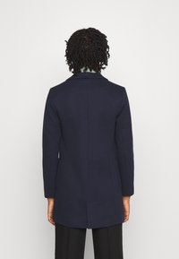 Námořnický modrý přizpůsobený kabát s klopami, dlouhými rukávy a dvěma bočními kapsami. Rovný střih a jemná vlněná textura.