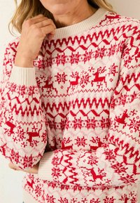 Person in einem cremefarbenen und roten Strickpullover mit Rentier- und Schneeflockenmuster, Hand nahe des Gesichts, oberer Teil des Körpers teilweise sichtbar.