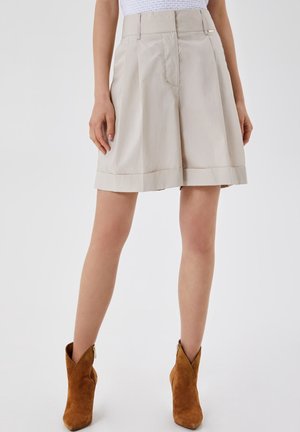 Shorts plissés taille haute beige portés avec des bottines en daim marron sur un mannequin debout devant un fond uni.