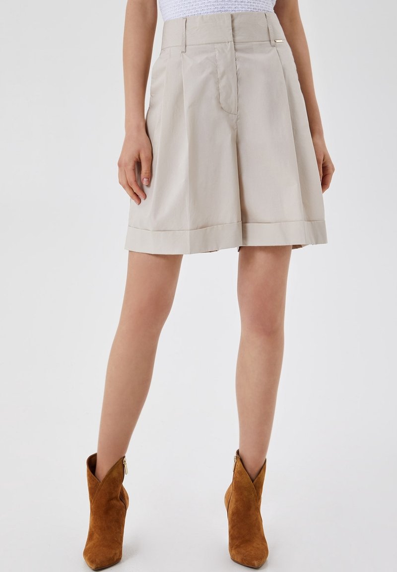 Shorts plissés taille haute beige portés avec des bottines en daim marron sur un mannequin debout devant un fond uni.