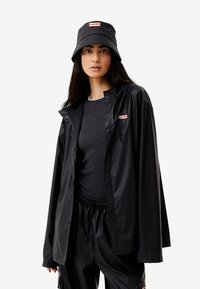 Svart vattentät jacka med tryckknappar, matchande svart bucket hat och grå tröja under. Har "HUNTER"-logotypen på båda plagg.