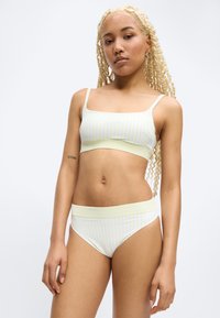 Νεαρή γυναίκα με μακριά ξανθά κοτσίδια, φορώντας ανοιχτού κίτρινου και λευκού ριγέ σουτιέν και σλιπ, όρθια μπροστά σε απλό λευκό φόντο.