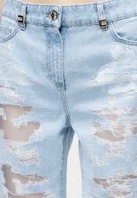 Svetlomodré riflové nohavice s veľkými rozstrapkanými časťami odhaľujúcimi priehľadnú látku pod nimi, strieborné zapínanie na gombík a dekoratívne kovové očká na opasok.